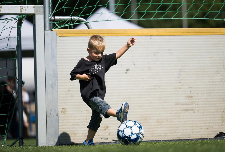 Ein kleiner Junge in einem schwarzen T-Shirt tritt auf einen Fußball vor einem Tor auf einem Rasenplatz.